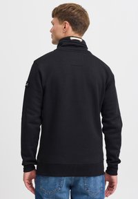 Sudadera negra con cuello alto, tejido texturizado y puños de canalé. Presenta un pequeño logotipo bordado en la manga y un detalle de cuerda en el cuello.