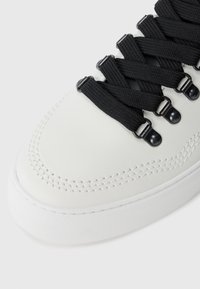 Hvite sneakers med en glatt skinnoverdel, intrikate sidesømmer og svarte tekstilskolisser festet med metalliske detaljer.
