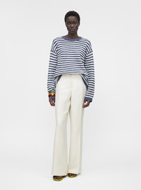 Femme portant un pull rayé bleu et blanc, un pantalon blanc taille haute à jambes larges, des sandales jaunes et des bracelets colorés, debout devant un fond uni.
