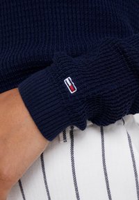Ingrandimento di un polsino di un maglione lavorato a maglia blu navy con un piccolo logo rettangolare rosso, bianco e blu sopra una mano appoggiata su pantaloni bianchi con righe verticali.