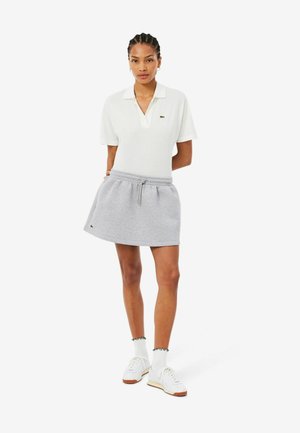Femme debout portant un polo blanc à manches courtes, une jupe grise avec cordon, des chaussettes blanches et des baskets blanches sur fond blanc.