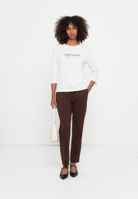 Witte shirt met driekwartmouwen met "TOM TAILOR" opdruk, gecombineerd met bruine taps toelopende broek en zwarte platte schoenen. Accessoires zijn onder andere een kleine lichte handtas.