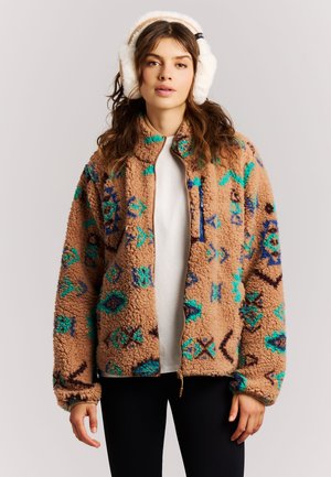 Femme portant une veste polaire beige à motif, chemise blanche, pantalon noir et cache-oreilles blancs et duveteux, debout devant un fond clair uni.
