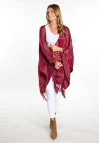 Burgundin poncho, jossa on geometrisia kuvioita ja reunoissa frillejä, valmistettu teksturoidusta kankaasta, päällä valkoinen toppi ja parina valkoiset housut.