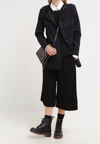 Schwarzer karierten Blazer mit taillierter Silhouette, über einem schwarzen Pullover und weiten schwarzen Culottes getragen. Schwarze Schnürstiefel mit gelben Akzenten.