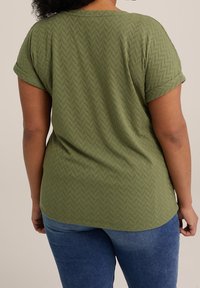 Olivgrön kortärmad topp med ett texturerat fiskbensmönster, med rund halsringning och en avslappnad passform. Bärs med blå jeans.