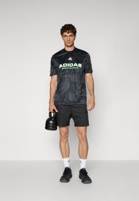 Black athletic t-shirt with green "ADIDAS SPORTSWEAR" logo, patterned shorts, and black shoes, holding a kettlebell in one hand.