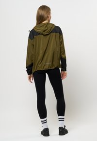 Giacca leggera verde oliva con dettagli neri, orlo con coulisse e cappuccio. Abbinata a leggings neri e calze sportive a righe.