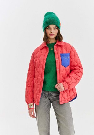 Młoda kobieta w zielonej czapce typu beanie, zielonym swetrze, koralowej pikowanej kurtce z niebieską kieszenią i szarych dżinsach, stojąca na tle jednolitego tła.