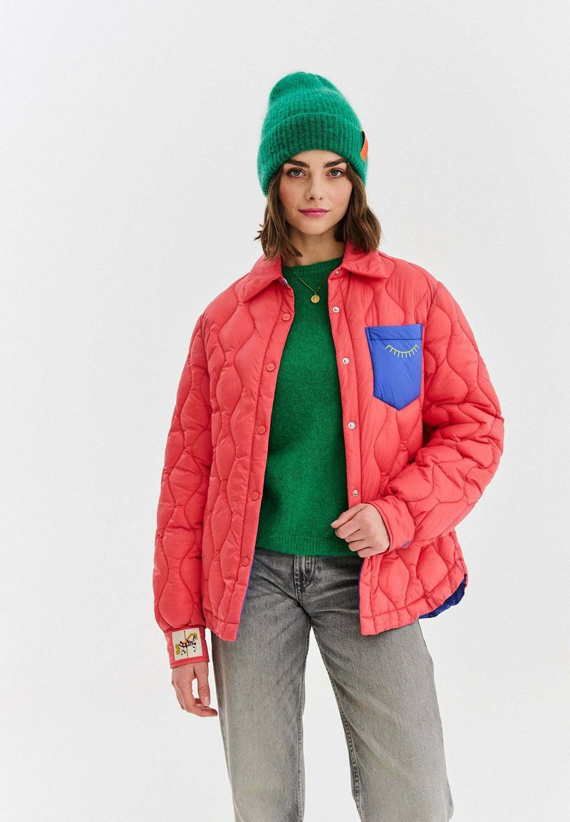 Mladá žena s zelenou čepicí, zeleným svetrem, korálově zbarvenou prošívanou bundou s modrým kapsou a šedými džíny, stojící před jednoduše pojatým pozadím.