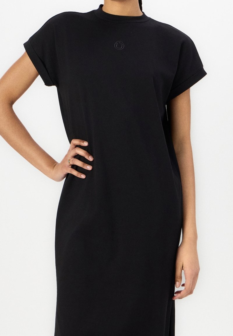 Femme portant une robe midi noire à manches courtes avec des poignets retroussés et un petit visage souriant brodé sur la poitrine.