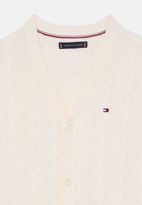 Crèmefarget cardigan laget av teksturert kabelstrikk, med V-hals, fem knapper og en liten brodert logo i rødt, hvitt og blått.
