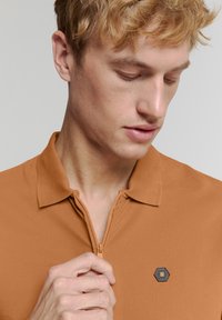 Polo shirt in oranjebruin stof met een ritskraag, gestructureerd oppervlak en een klein hexagonaal logo op de borst.