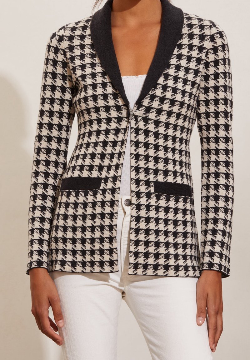 Mustavalkoinen houndstooth-bleiseri, jossa on tumma kaulus, kaksi etutaskua ja säädettävä muoto. Rakennepidempi kangas, jossa on jämäkkä siluetti.
