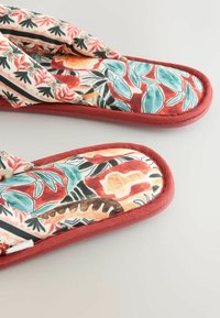 Minkšti audinio šlepetės su įvairiaspalviais gėlių ir lapų raštais raudonos, žalsvai mėlynos, oranžinės ir juodos spalvų, su rūdžių raudonos spalvos apvadais.