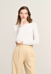 Long-sleeve white top with a boat neckline, paired with beige high-waisted trousers featuring pleats and a tailored fit. Smooth texture throughout.