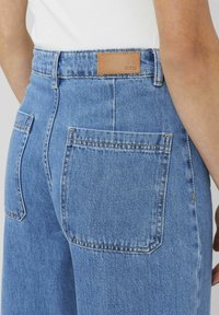 Jean en denim bleu à taille haute, avec deux poches arrière et une étiquette en cuir à la ceinture marquée "B2B". Texture lisse.