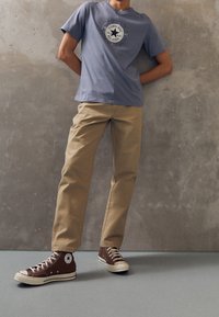 Light blue short-sleeve t-shirt with a circular black logo, tan pants, and brown high-top sneakers with white accents and a star logo.