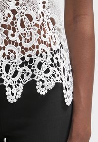 Top in pizzo bianco con intricati motivi floreali e design a trama aperta, caratterizzato da un orlo scalloppato. Indossato su un tessuto nero.