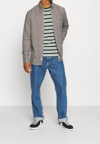 Bruine geruite overhemd over een groen-wit gestreept T-shirt, gecombineerd met opgerolde blauwe jeans en zwarte sneakers met witte zolen.