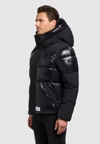 Čierna prešívaná bunda s lesklým povrchom, vysokým golierom, kapucňou, vreckami na zips a štítkom na spodnom okraji. Textúrovaný materiál.