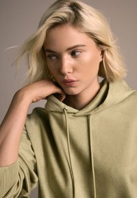 Olivovo zelená mikina s jemnou textúrou, klokanie vrecko, nastaviteľná kapucňa so šnúrkami a rebrované manžety. Modelka má ruku opretú o bradu.