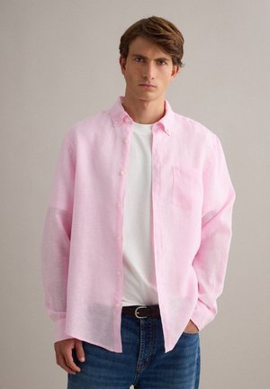 Jonge man met bruin haar die een open lichtroze overhemd draagt over een wit T-shirt en blauwe spijkerbroek tegen een effen achtergrond.