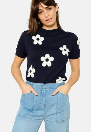 Maglione corto a maniche corte di colore blu navy con motivi floreali bianchi; abbinato a pantaloni in denim chiaro a vita alta con tasche frontali.