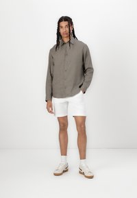 Camisa de lino ligera color caqui con cierre de botones, combinada con pantalones cortos blancos y zapatillas beige con detalles de tela y suelas de goma.