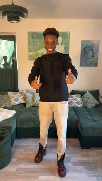 Nuori mies hymyilee ja osoittaa eteenpäin sormipistoilla, seisten olohuoneessa, jossa vihreä sohva ja seinätaide hänen takanaan.