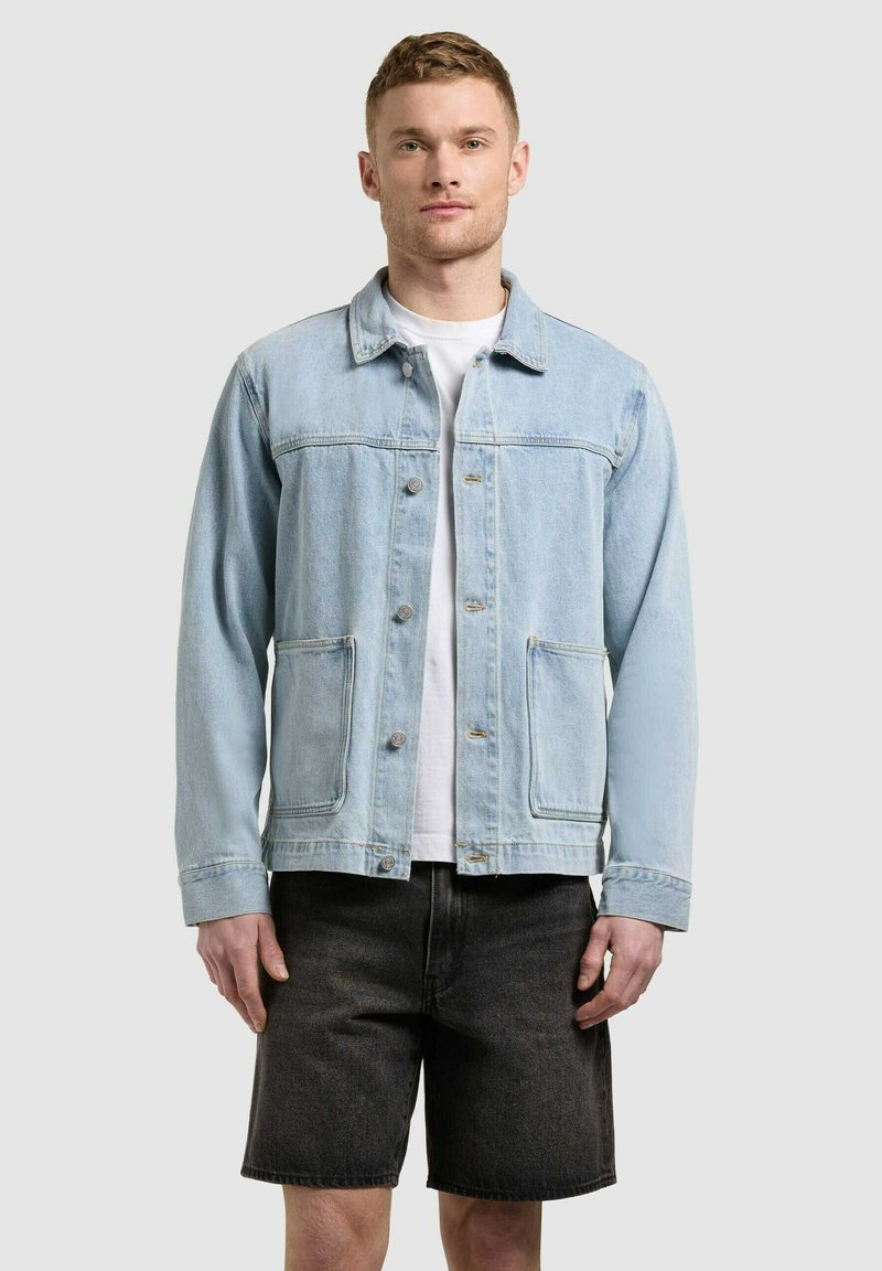 Homme portant une veste en jean bleu clair sur une chemise blanche et un short en jean gris foncé, debout devant un fond uni clair.