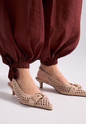 Pies con zapatos beige de tacón bajo y tira trasera, con patrones recortados y detalle de hebilla dorada, combinados con pantalones burdeos sueltos y fruncidos.