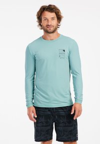 Ilgapirštis, mėtų žalios spalvos rash guard'as su grioveliais ir juoda grafine kišene. Derinamas su juodais maudymosi šortais, puikuojančiais palmės medžių raštu.