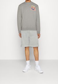 Felpa grigia con design grafico sul petto, abbinata a pantaloni corti grigi chiari e sneakers bianche, con vestibilità rilassata e polsini.