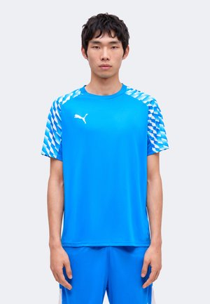 Tânăr în picioare cu fața înainte, purtând un tricou sport Puma albastru deschis cu mâneci cu model și pantaloni scurți albaștri asortați.