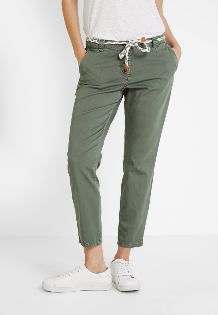 Pantalon kaki, coupe décontractée avec taille à cordon, poches latérales et accents blancs. Porté avec des baskets blanches. Tissu en coton lisse.