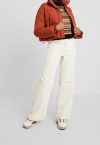 Ruskioranssi pehmustettu takki korkealla kauluksella, yhdistettynä kuviolliseen neulepaitaan. Kermanväriset vakosamettiset leveälahkeiset housut ja massiiviset valkoiset lenkkarit.