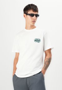 Witte katoenen t-shirt met korte mouwen, voorzien van een groene grafische afbeelding op de linkerborst. Het model draagt zwarte zonnebril en grijze broek.