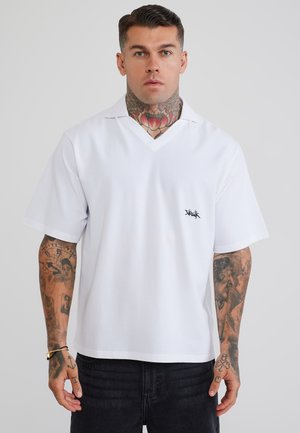 Mann mit Hals- und Arm-Tattoos trägt ein weißes kurzärmeliges Hemd mit einem kleinen schwarzen Logo auf der Brust und schwarze Hosen vor einem schlichten Hintergrund.