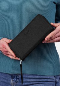Black fabric wallet with a zipper closure, featuring a branded emblem embossed on the front, held against a ribbed teal sweater.