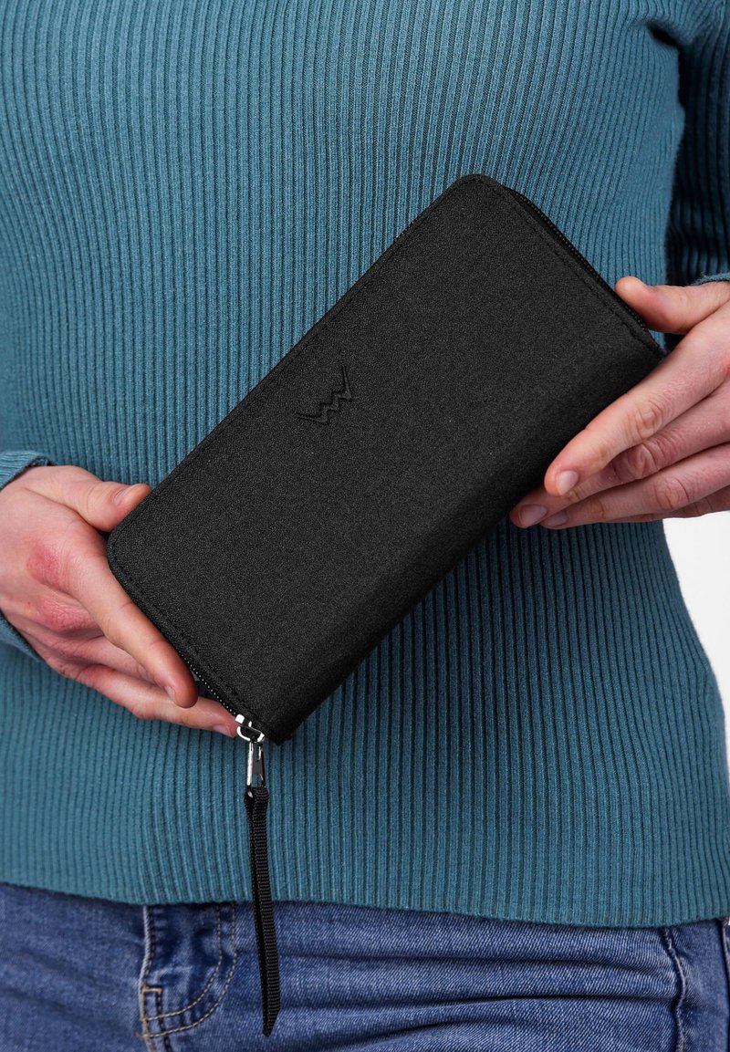 Black fabric wallet with a zipper closure, featuring a branded emblem embossed on the front, held against a ribbed teal sweater.