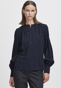 Navyblauwe blouse met knoopsluiting en geplooide halslijn. Kenmerken zijn lange, volumineuze mouwen en een gladde, gestructureerde stof.