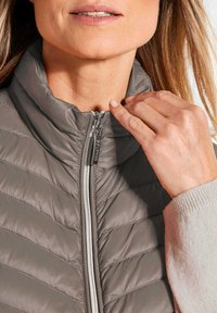 Hellgraue gesteppte Weste mit hohem Kragen und Reißverschluss. Der Stoff wirkt glatt und hat diagonale Steppmuster sowie silberne Hardware.