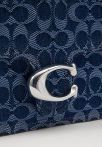 Denim bag with an embossed pattern of linked circles in dark blue. Silver metal hardware clasp, outlined in contrasting brown stitching.