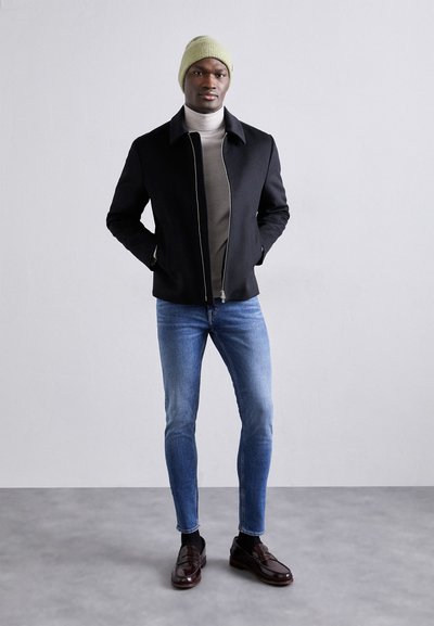 Melnā jaku ar rāvējslēdzēju un augstu apkakli, pelēks turtlenecks, šauras zilas džinses un brūni loafers. Modelis nēsā gaiši zaļu adītu beanie cepuri.