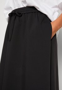 Personne portant un pantalon noir à cordon avec la main dans la poche et une chemise blanche rentrée visible à la taille.