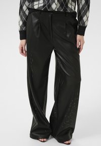 Fekete perforált nadrág széles szabással, redőkkel és sima textúrával. Egy argyle mintás toppal kombinálva.