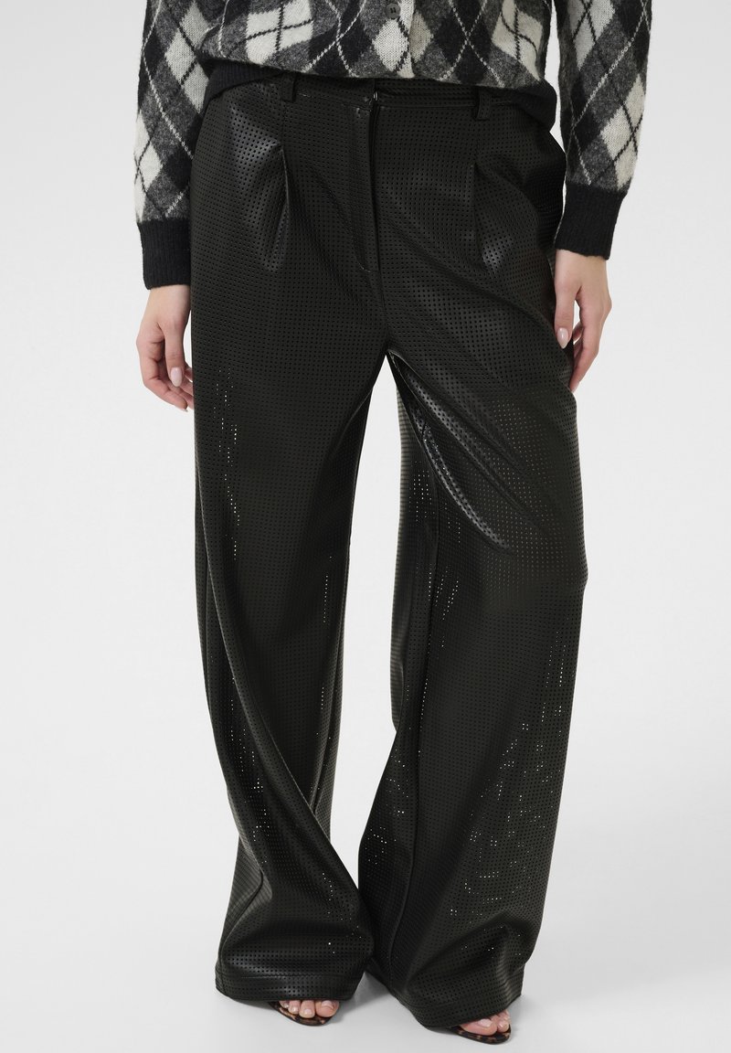 Pantalons noirs perforés avec une coupe loose, dotés de plis et d'une texture lisse. Associés à un haut à motif argyle.