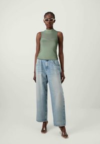 Top de tanque verde canelado combinado com jeans de ganga de perna larga em azul claro. Modelo usa óculos de sol transparentes e sandálias de salto transparentes.