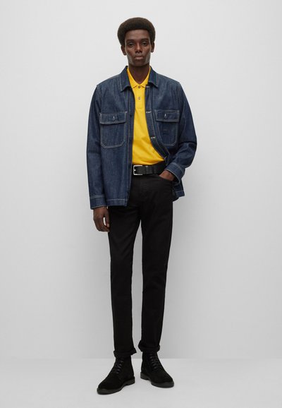 Veste en denim avec deux poches poitrine, polo jaune, pantalon noir et bottes noires. Le modèle se tient devant un fond clair.